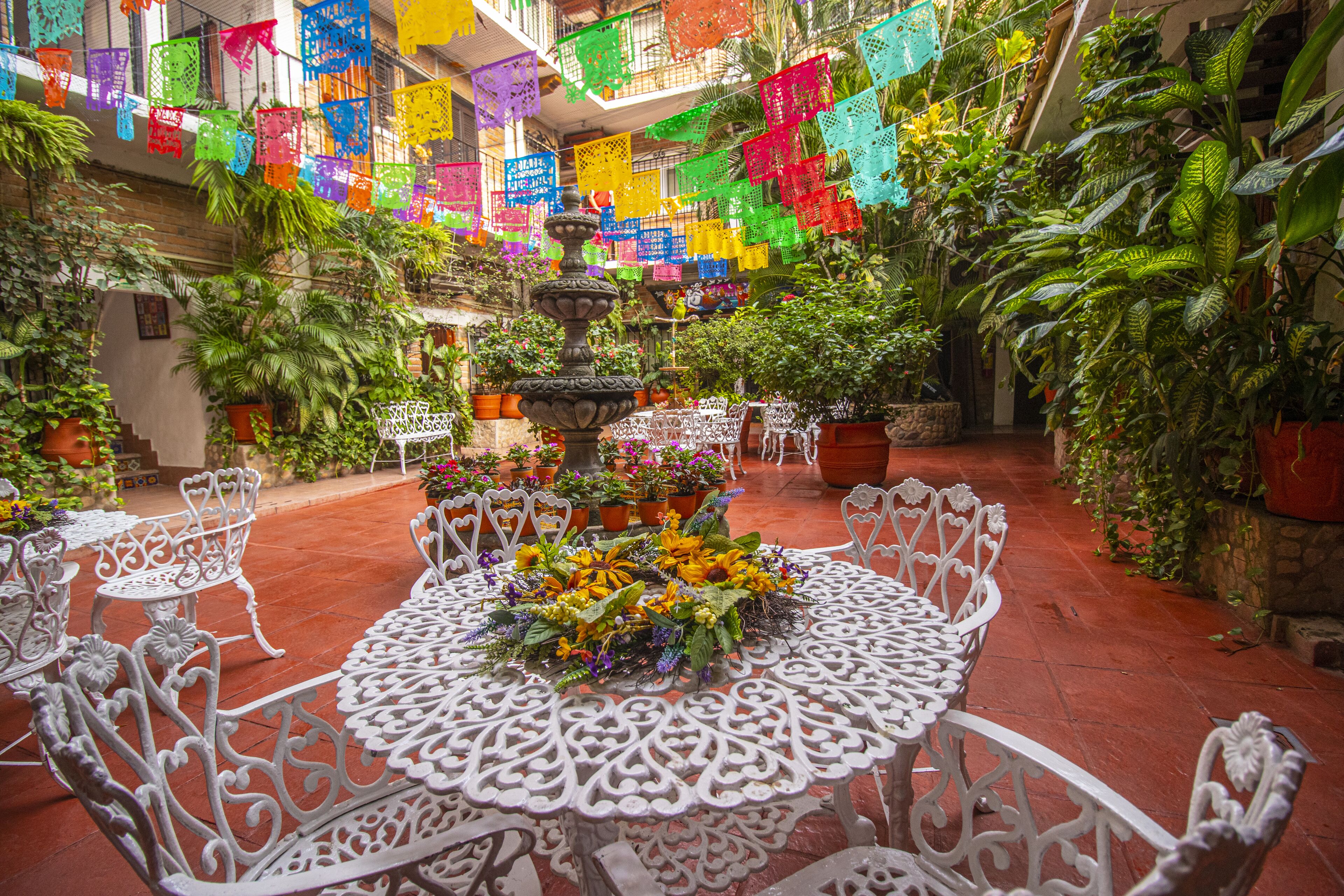 Un patio con una fuente, mesas y sillas blancas ornamentadas, rodeado de plantas en macetas y coloridas decoraciones colgantes.