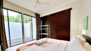 A hotel room with a bed, a ceiling fan, a window with a view of greenery, and a door.