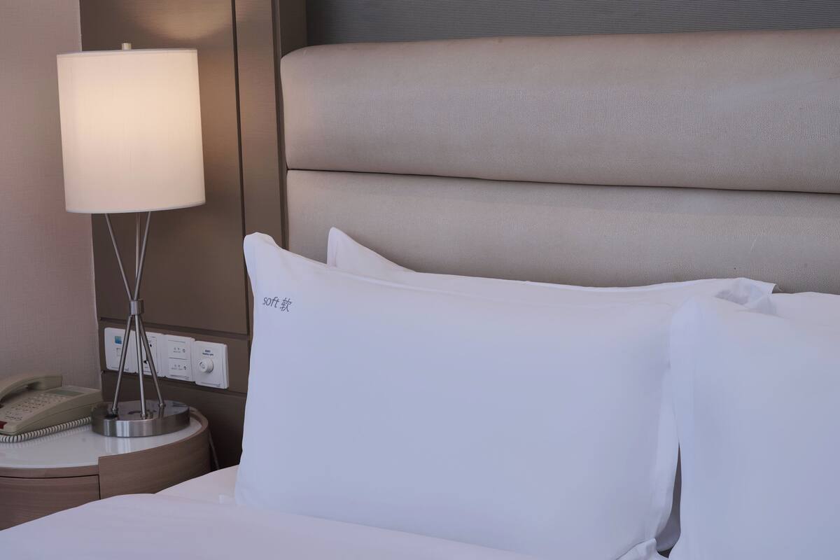 A hotel room with a bed, white pillows, a bedside lamp, a phone, and a control panel on the wall.