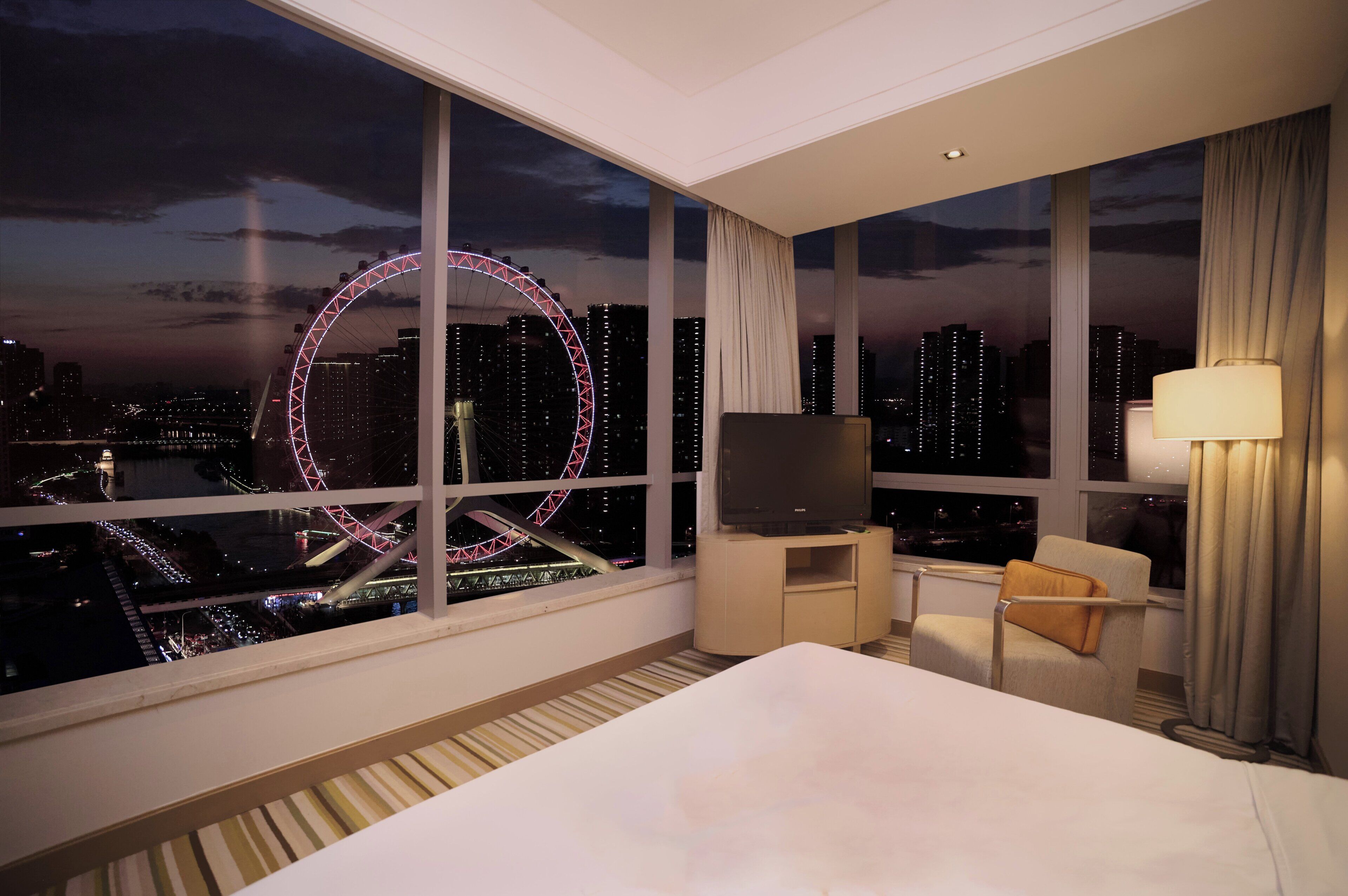 Una habitación de hotel con un ventanal grande, una cama, un televisor sobre un soporte, una silla y una lámpara.