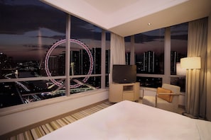 Una habitación de hotel con un ventanal grande, una cama, un televisor sobre un soporte, una silla y una lámpara.