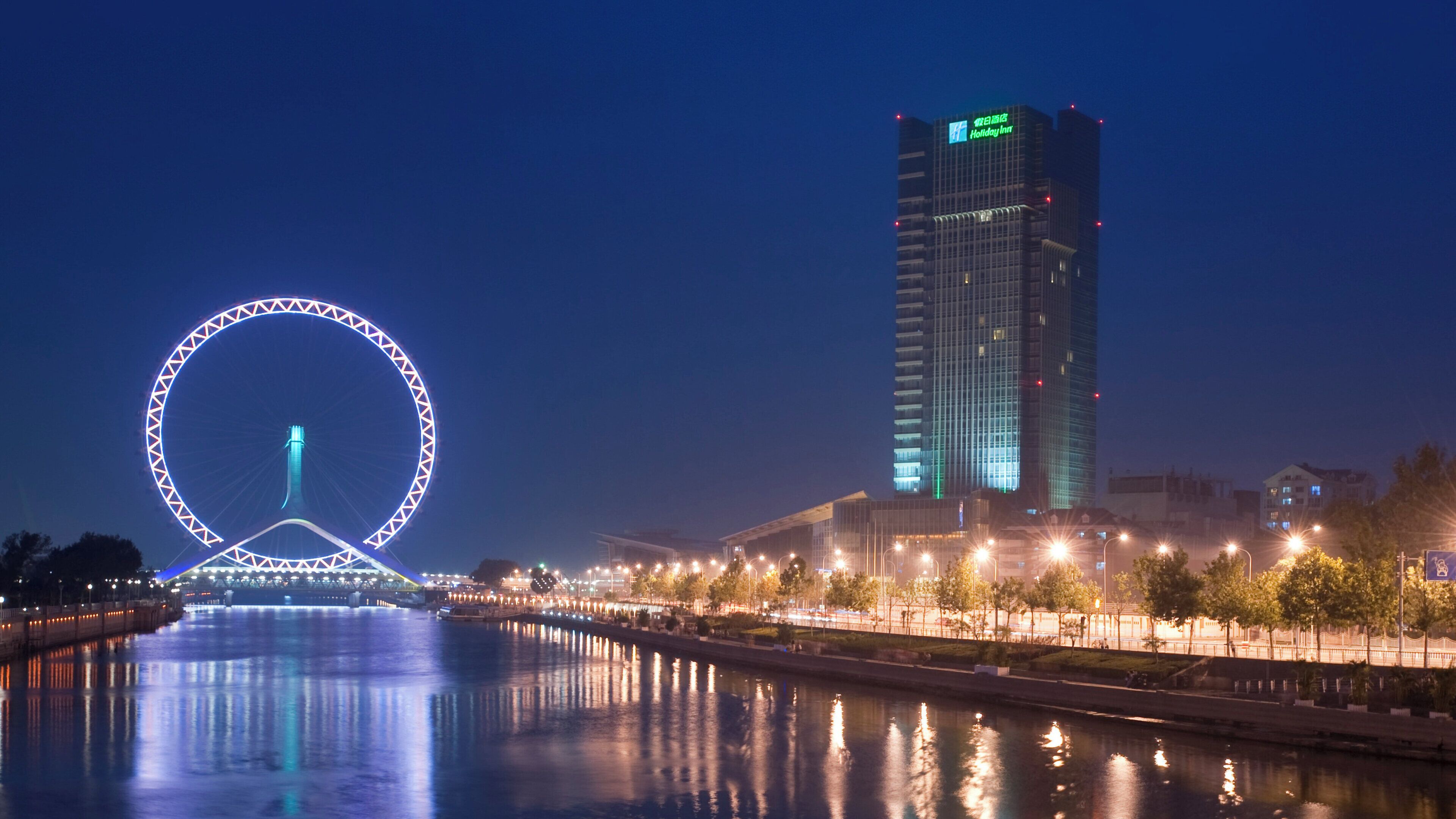 A waterfront view with a large illuminated Ferris wheel and a modern high-rise hotel.