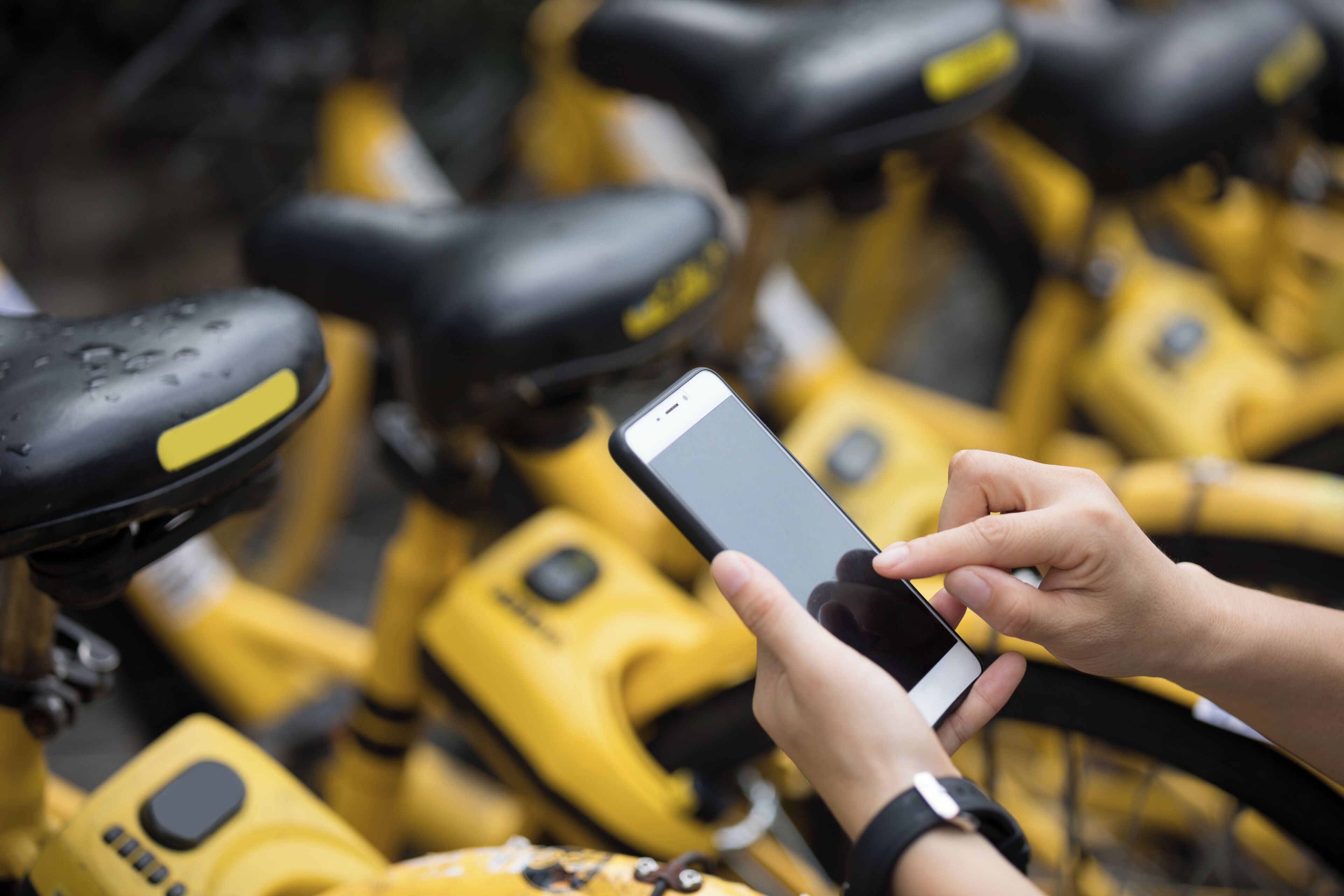A person holding a smartphone, possibly using a bike-sharing app, with yellow bicycles in the background.