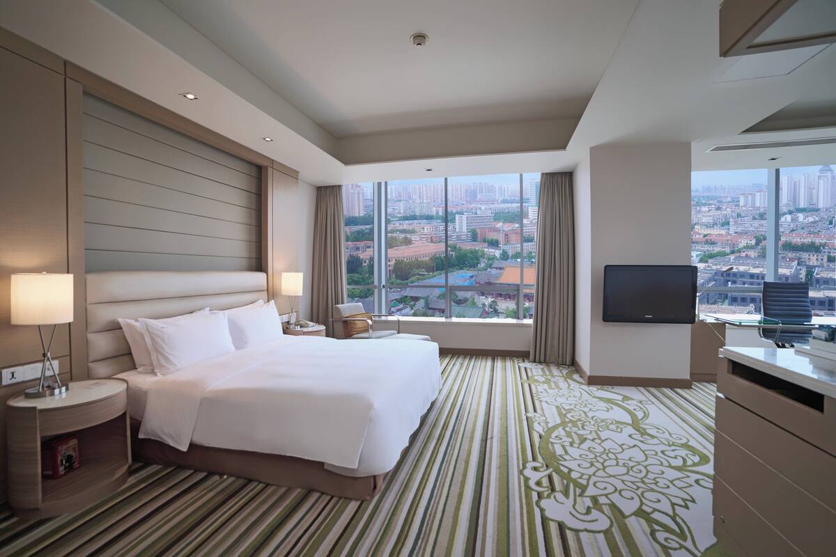 A hotel room with a large bed, a TV, a desk, and a view of the city.