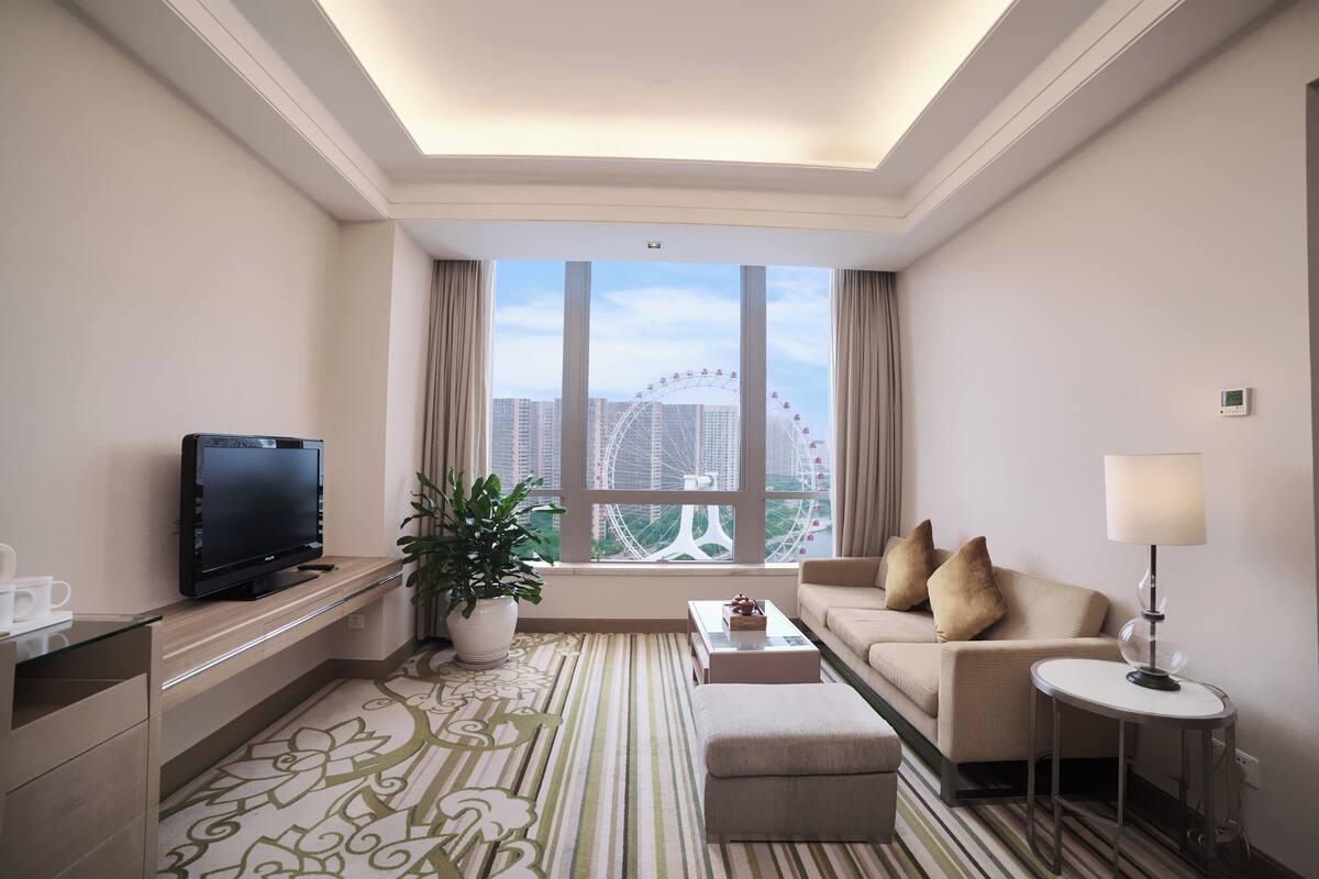 A hotel room with a large window, a flat-screen TV, a sofa, a coffee table, a lamp, and a potted plant.