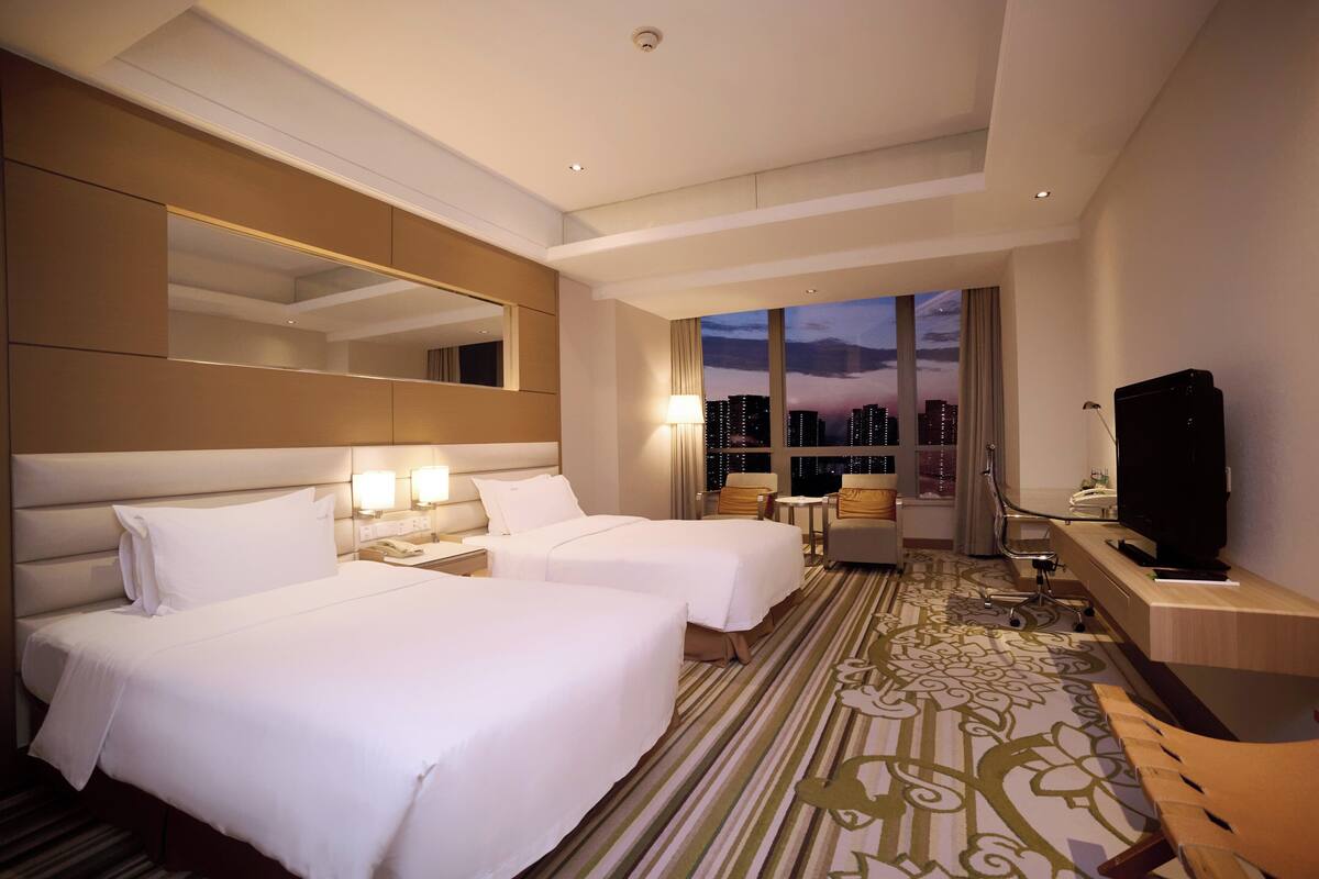 A hotel room with two beds, a large mirror, a television, and a view of the cityscape at dusk.
