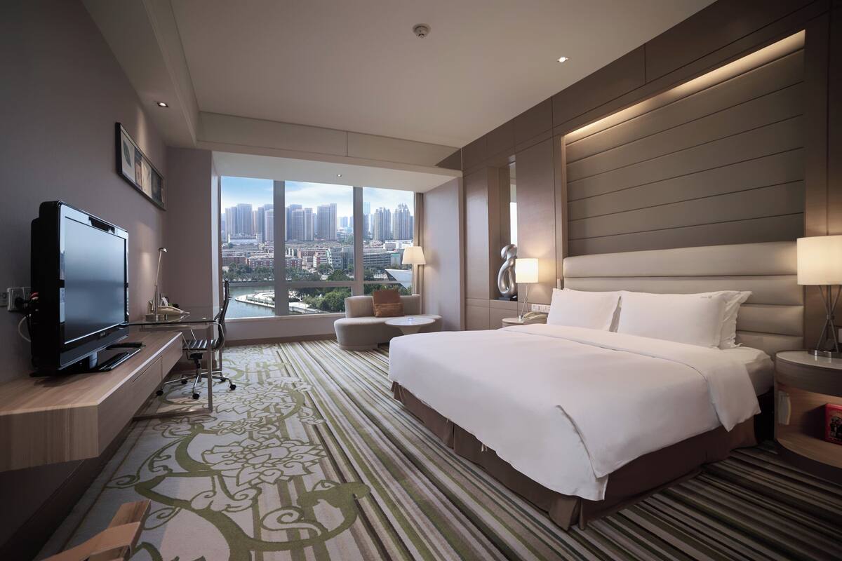 A modern hotel room with a large bed, a TV, a sofa, and a view of the cityscape.
