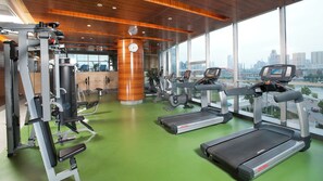 Ein Fitnessstudio mit Trainingsgeräten, einer großen Uhr und Blick auf eine Stadtlandschaft durch hohe Fenster.