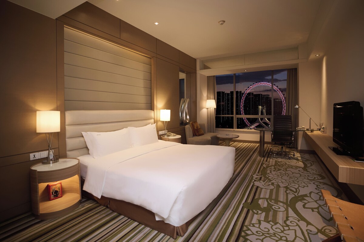 A hotel room with a large bed, a desk, a TV, and a view of a Ferris wheel and cityscape.