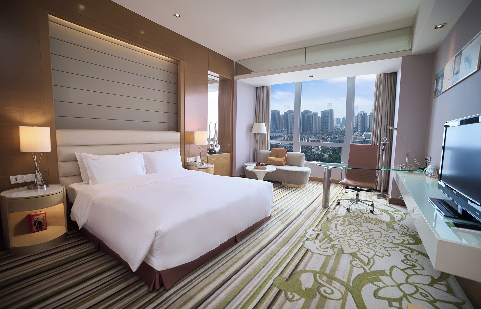 A hotel room with a large bed, a desk with a TV, a sofa, and a view of a cityscape.