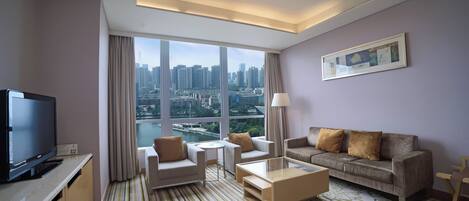 A modern hotel room with a large window offering a city view, a flat-screen TV, a sofa, a coffee table, and a lamp.