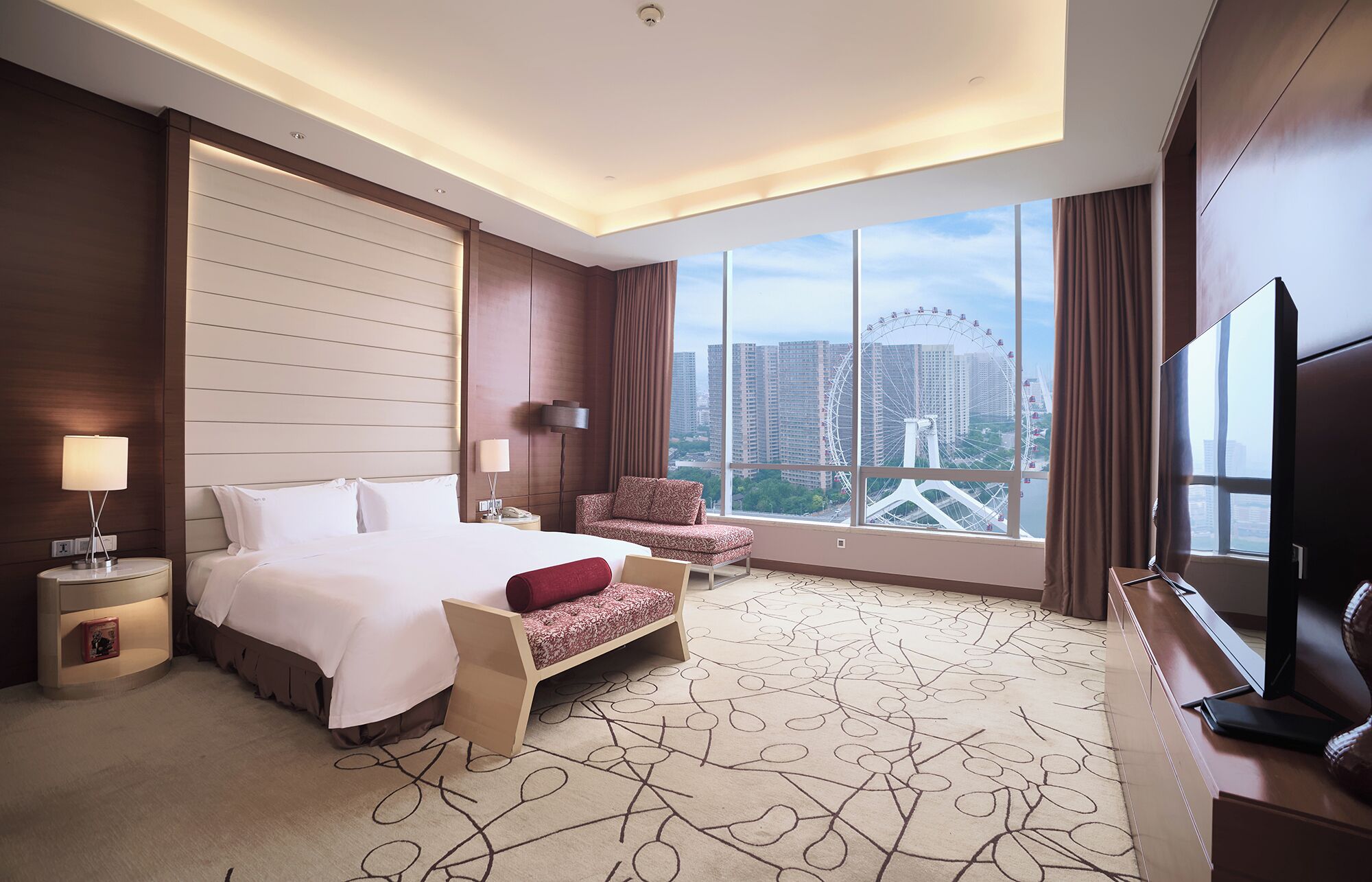 A hotel room with a large bed, a desk with a TV, a chair, and a view of a Ferris wheel.