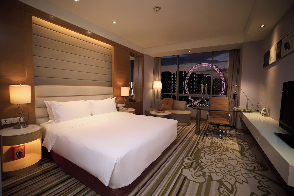 Quarto de hotel com uma cama grande, uma escrivaninha, uma cadeira, uma televisão e vista para uma roda gigante à noite.