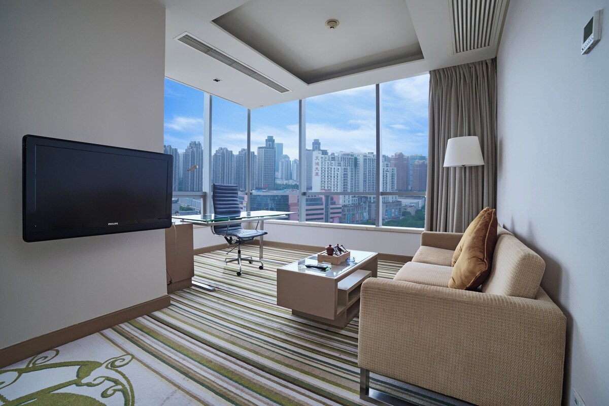 A modern hotel room with a large window offering a city view, a flat-screen TV, a sofa, a coffee table, and a lamp.