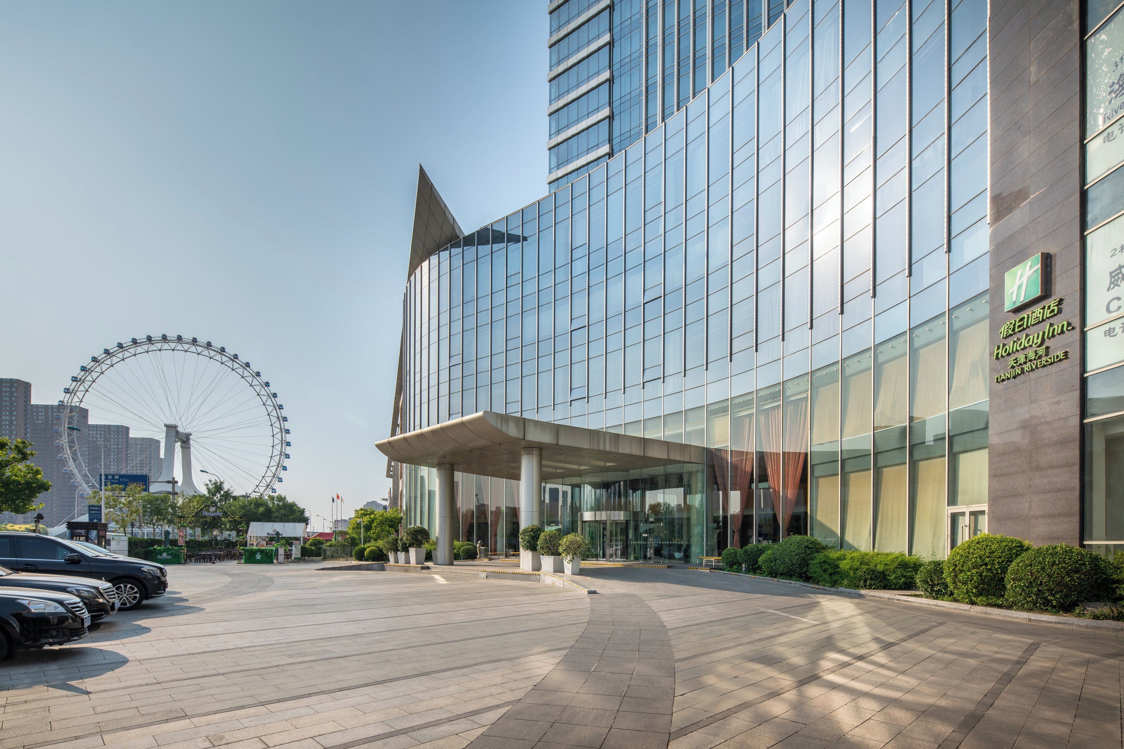 A modern hotel with a glass facade, a covered entrance, and a parking lot with cars.