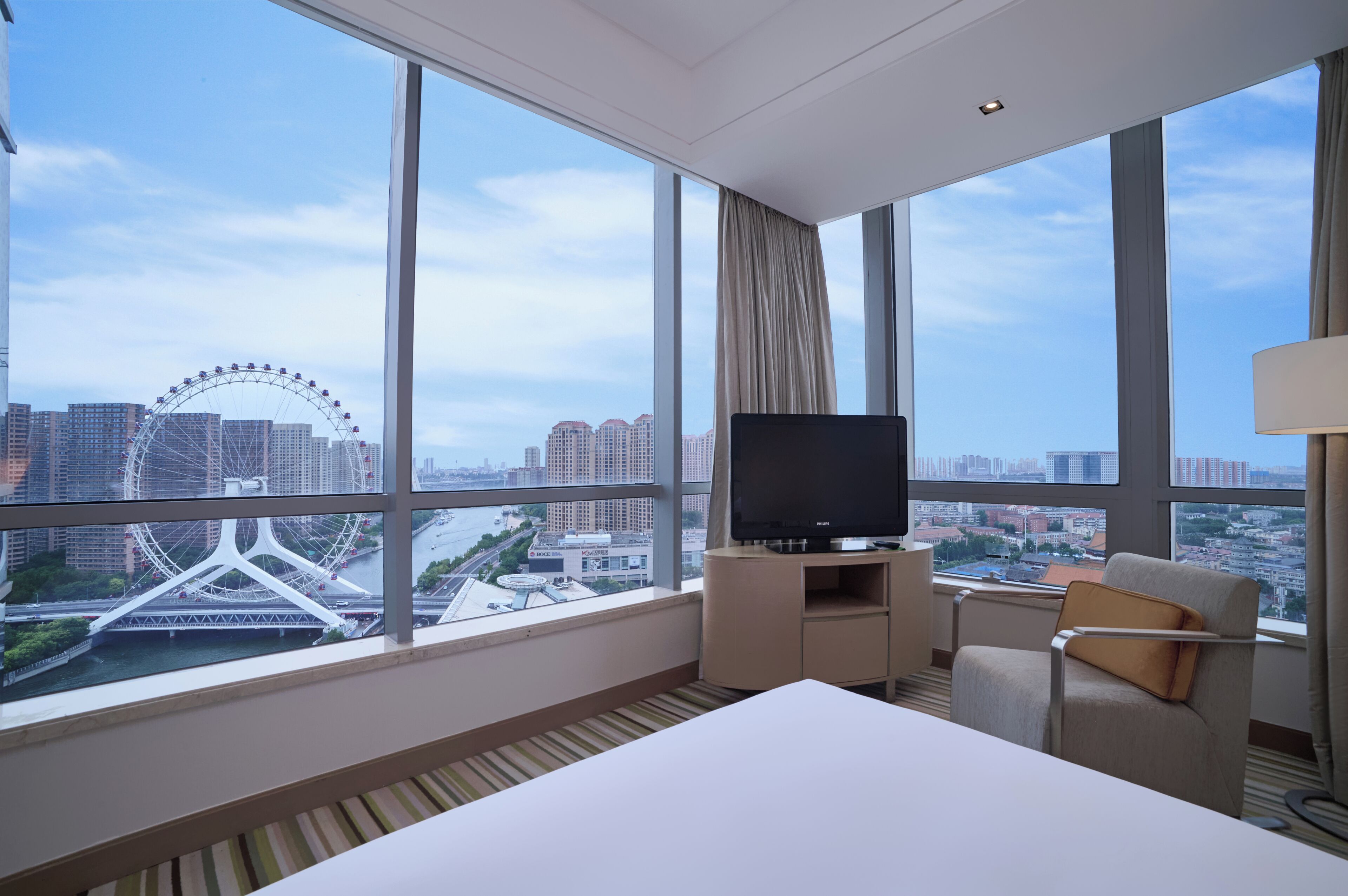 Habitación de hotel con un ventanal grande, un televisor de pantalla plana, una cama, una silla y vistas a un paisaje urbano que incluyen un puente y una noria.