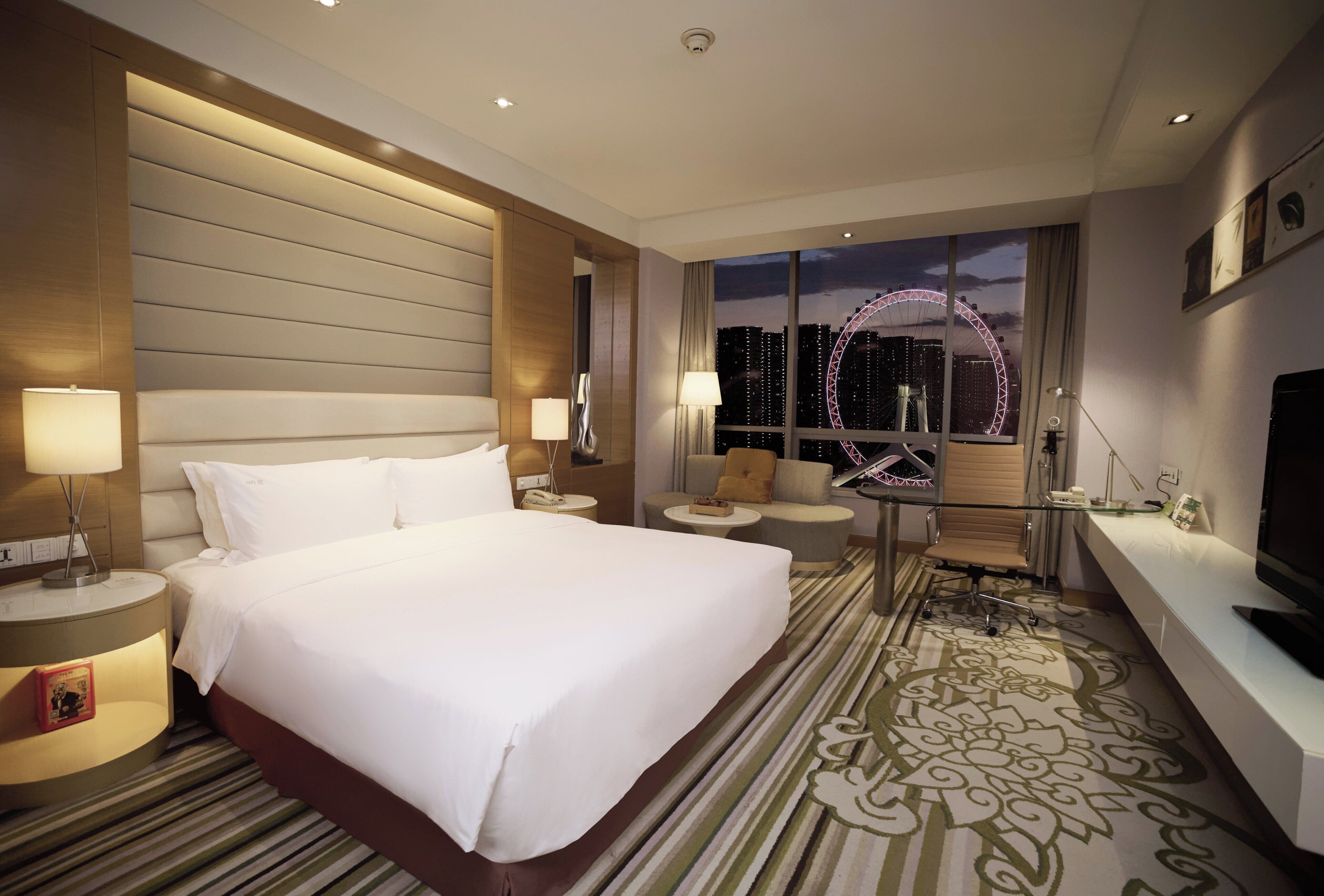 A hotel room with a large bed, a desk, a chair, a TV, and a view of a Ferris wheel at night.