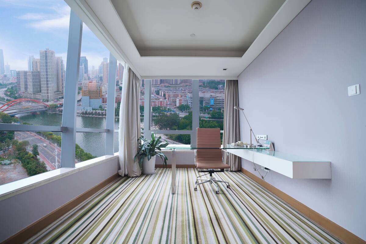 A modern hotel room with a large window offering a city view, a striped floor, a desk with a lamp, and a potted plant.