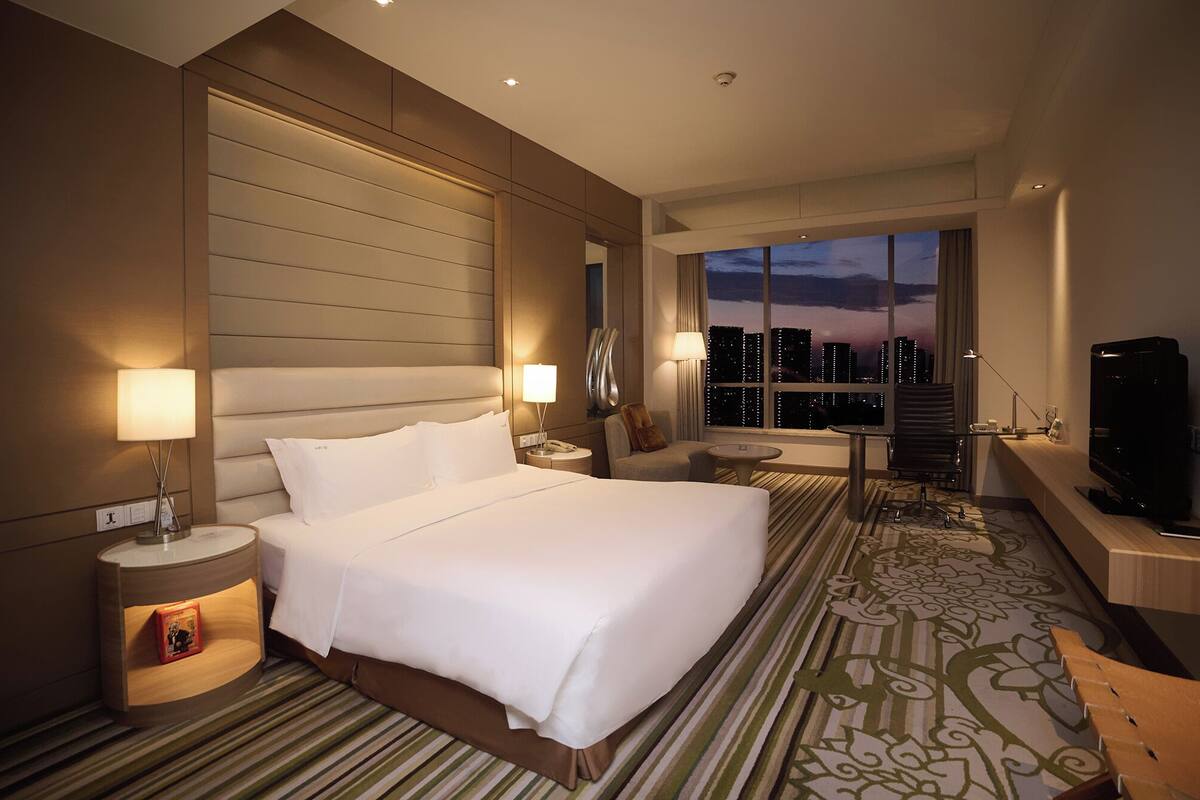 Habitación de hotel con una cama grande, un escritorio, un televisor y vistas a la ciudad al anochecer.