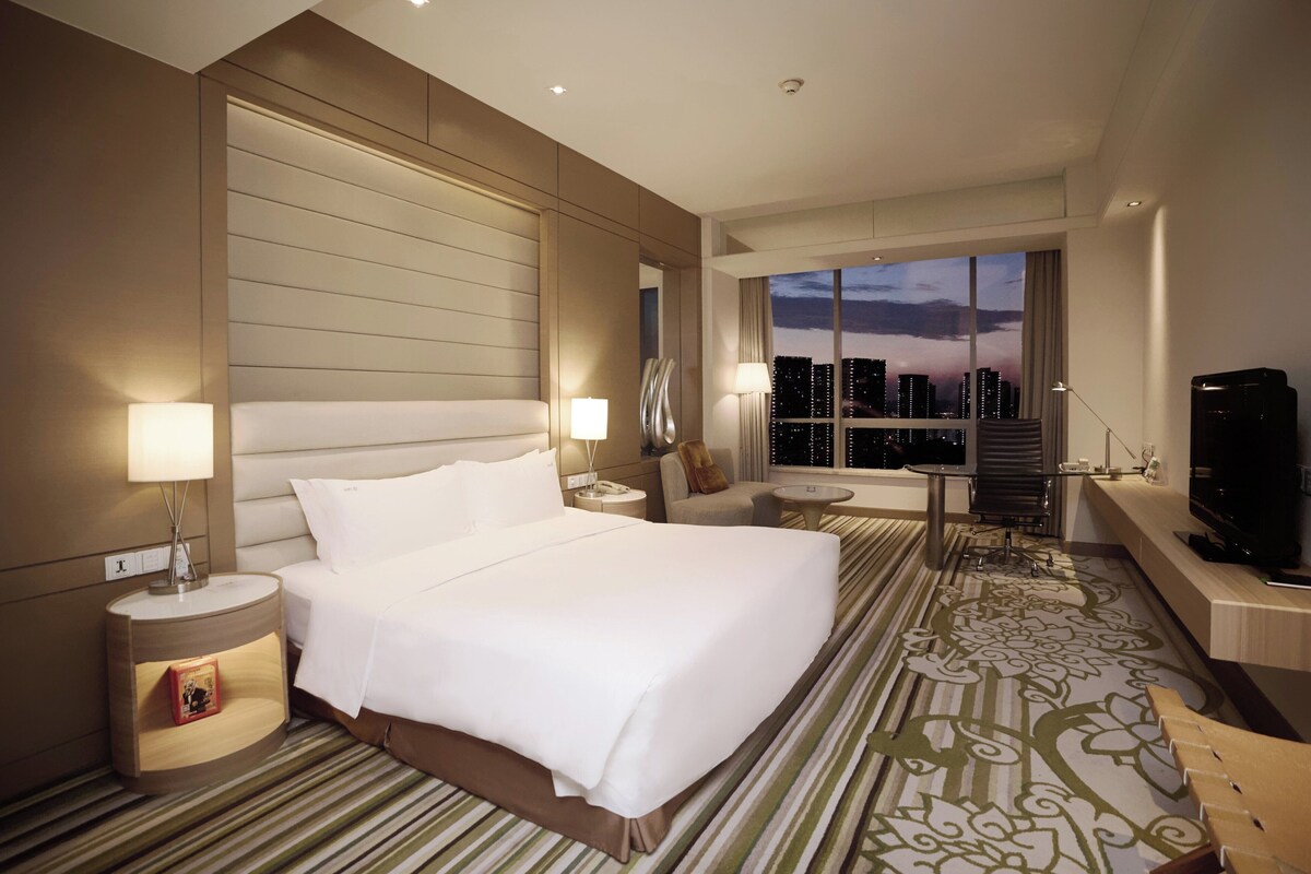 A hotel room with a large bed, a desk, a TV, and a view of the cityscape.