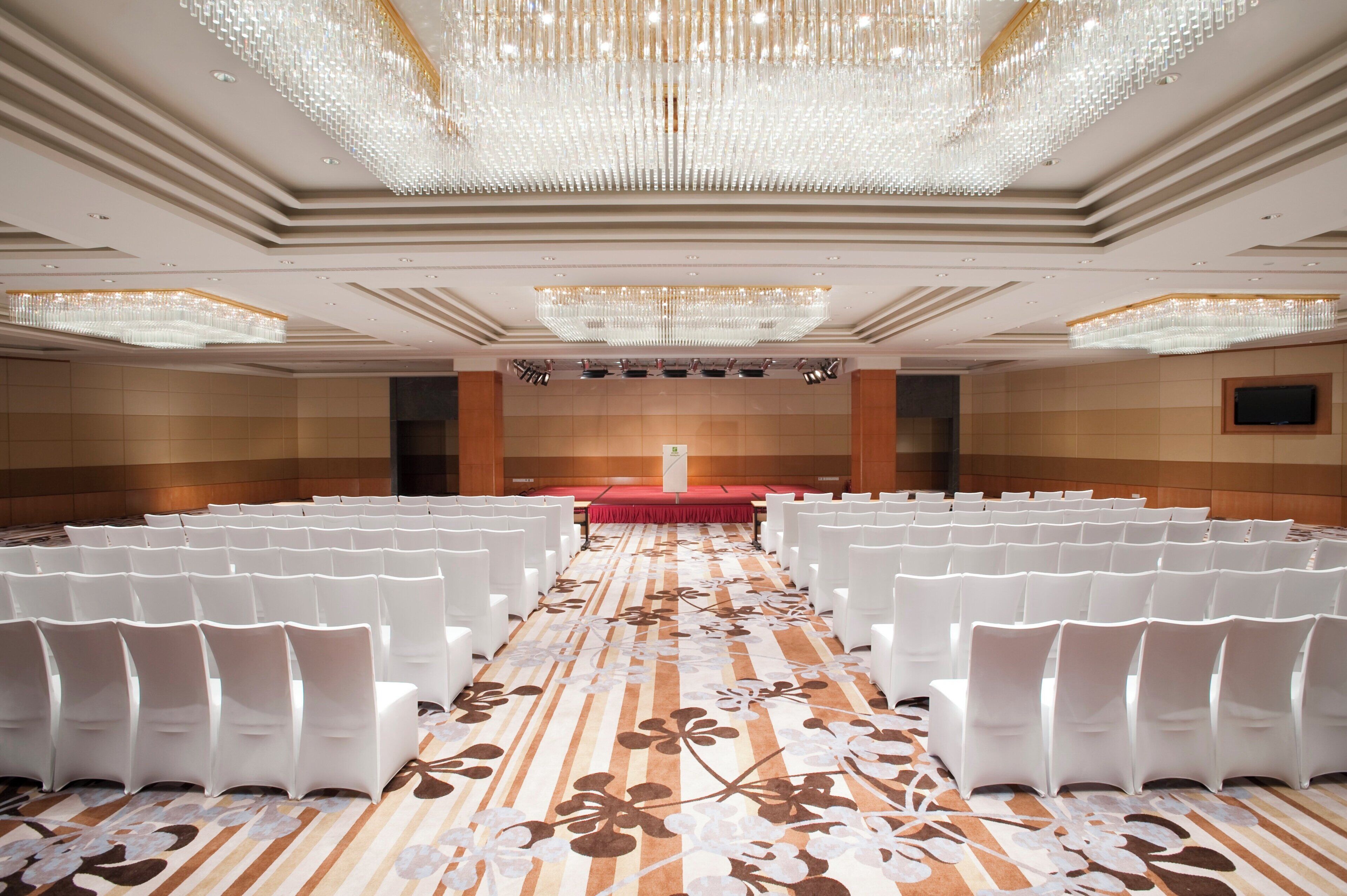 Un salón de conferencias con filas de sillas blancas, un escenario rojo y una gran araña.