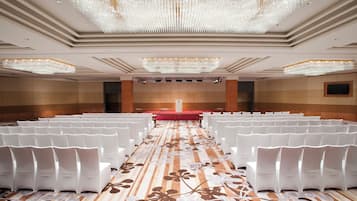 Un salón de conferencias con filas de sillas blancas, un escenario rojo y una gran araña.