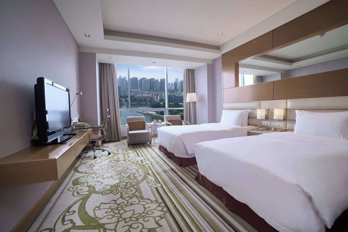 A hotel room with two beds, a TV, a desk, a chair, and a view of a cityscape.