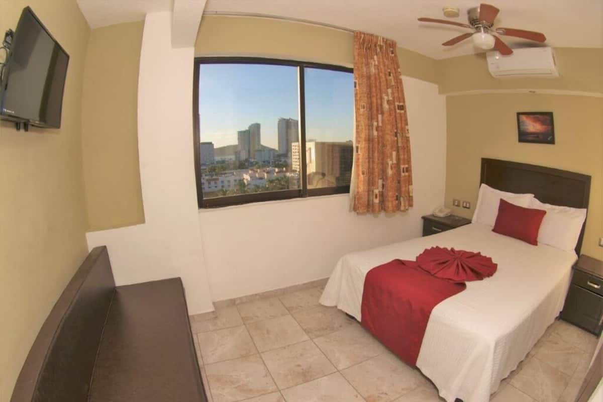 Habitación de hotel con cama, televisión, ventana con cortinas, ventilador de techo y vista a la ciudad.