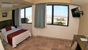A hotel room with a bed, a desk, a chair, a TV, and a window with a city view. - Hotel Playa Bonita (Mazatlán)
