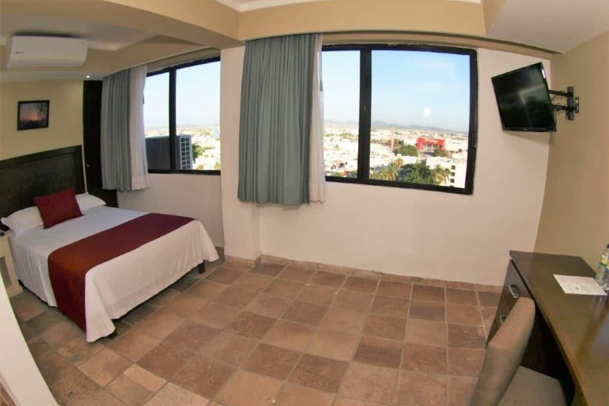 Habitación de hotel con cama, escritorio, silla, televisor y ventana con vista a la ciudad.