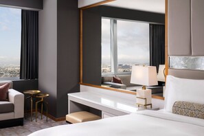 A modern hotel room with a large bed, a desk, a chair, a small table, and a large window offering a city view.