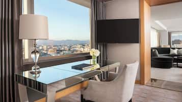 A modern hotel room with a glass desk, a comfortable chair, a flat-screen TV mounted on the wall, and a view of the cityscape through large windows.