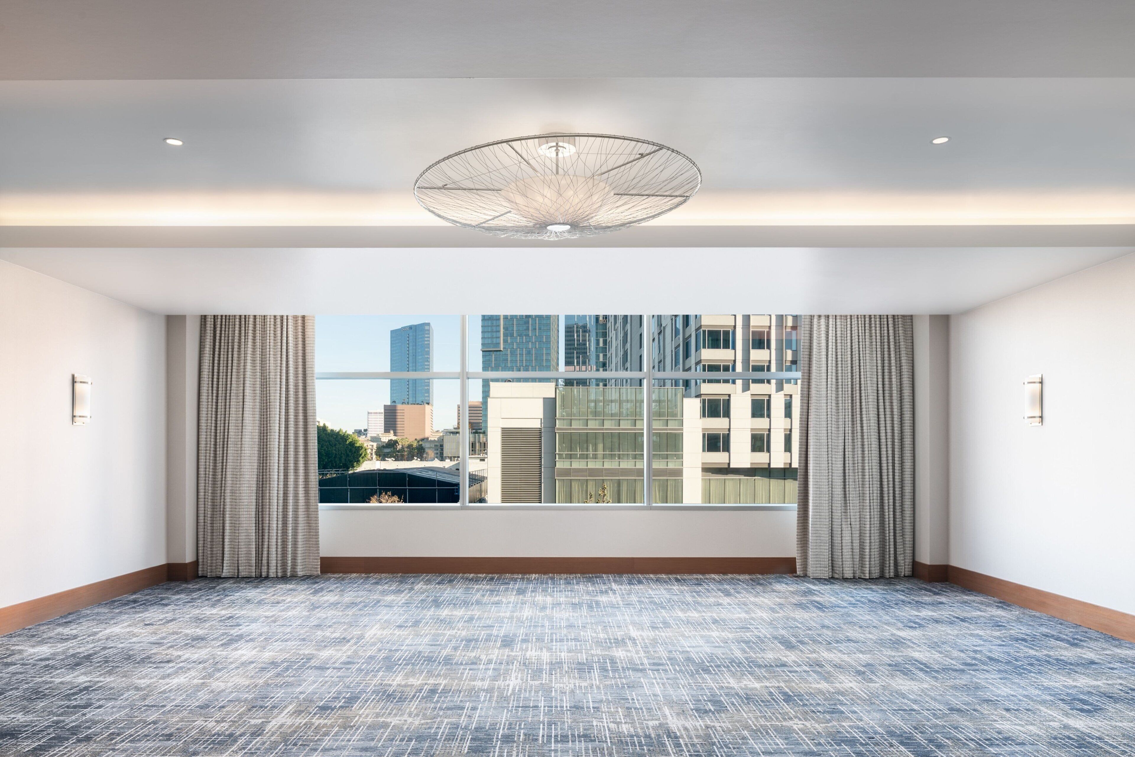 A spacious hotel room with a large window, a patterned carpet, and a modern ceiling light fixture.