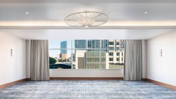 A spacious hotel room with a large window, a patterned carpet, and a modern ceiling light fixture.