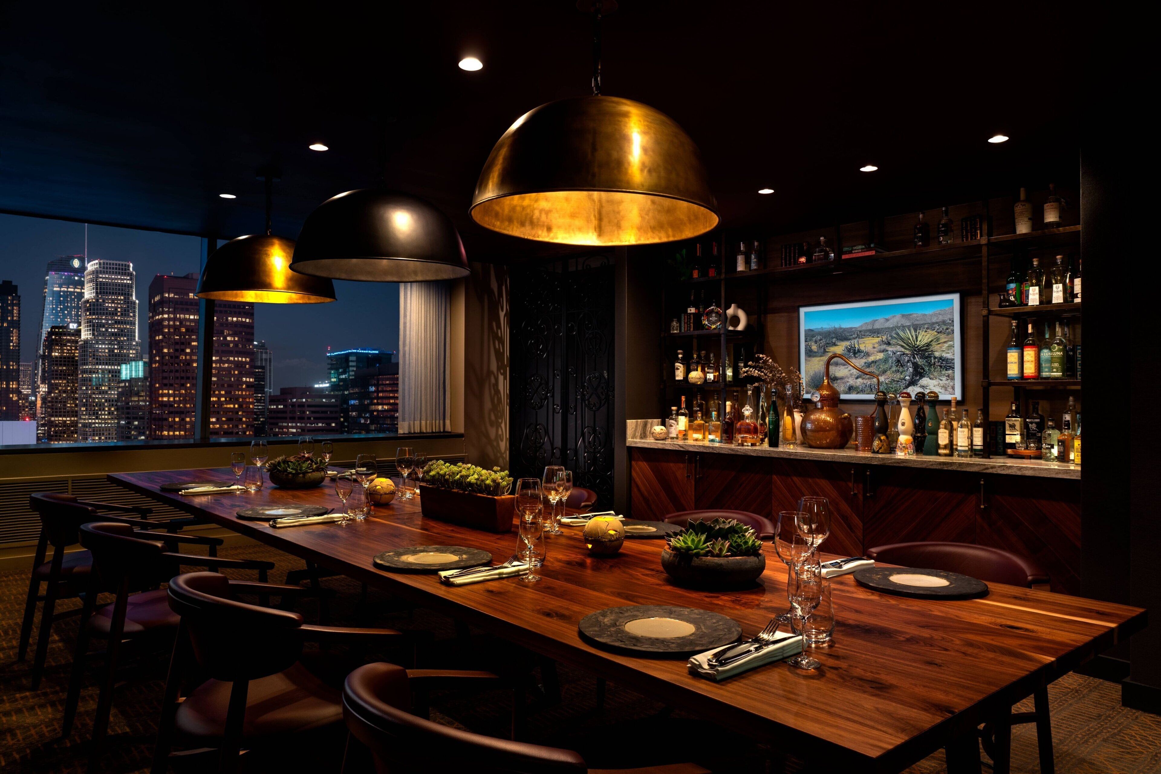 A restaurant with a city view, a long wooden table set for dining, and a bar area with bottles and glasses.
