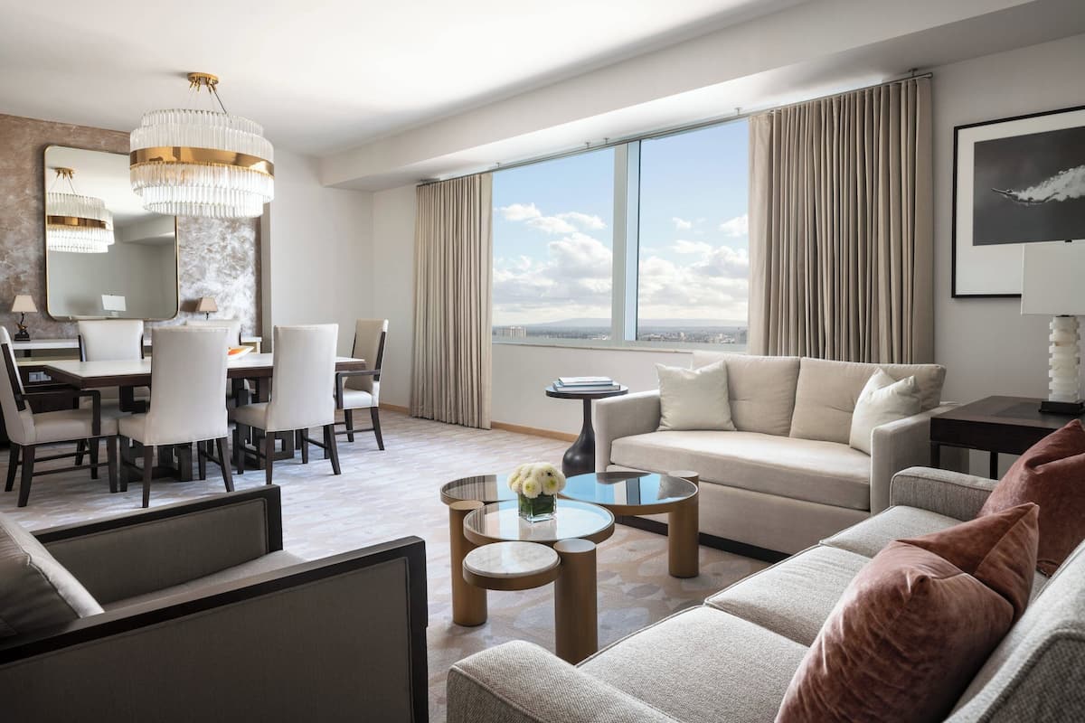 A modern hotel room with a dining area, a sofa, and a large window with a city view.