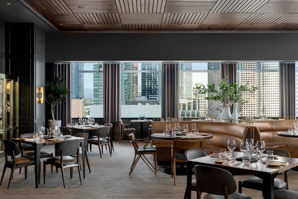 A modern restaurant with multiple seating areas, including tables and a sofa, with a city view through large windows.