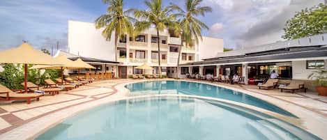 A pool area with lounge chairs and umbrellas, a building with balconies and palm trees, and a clear sky.