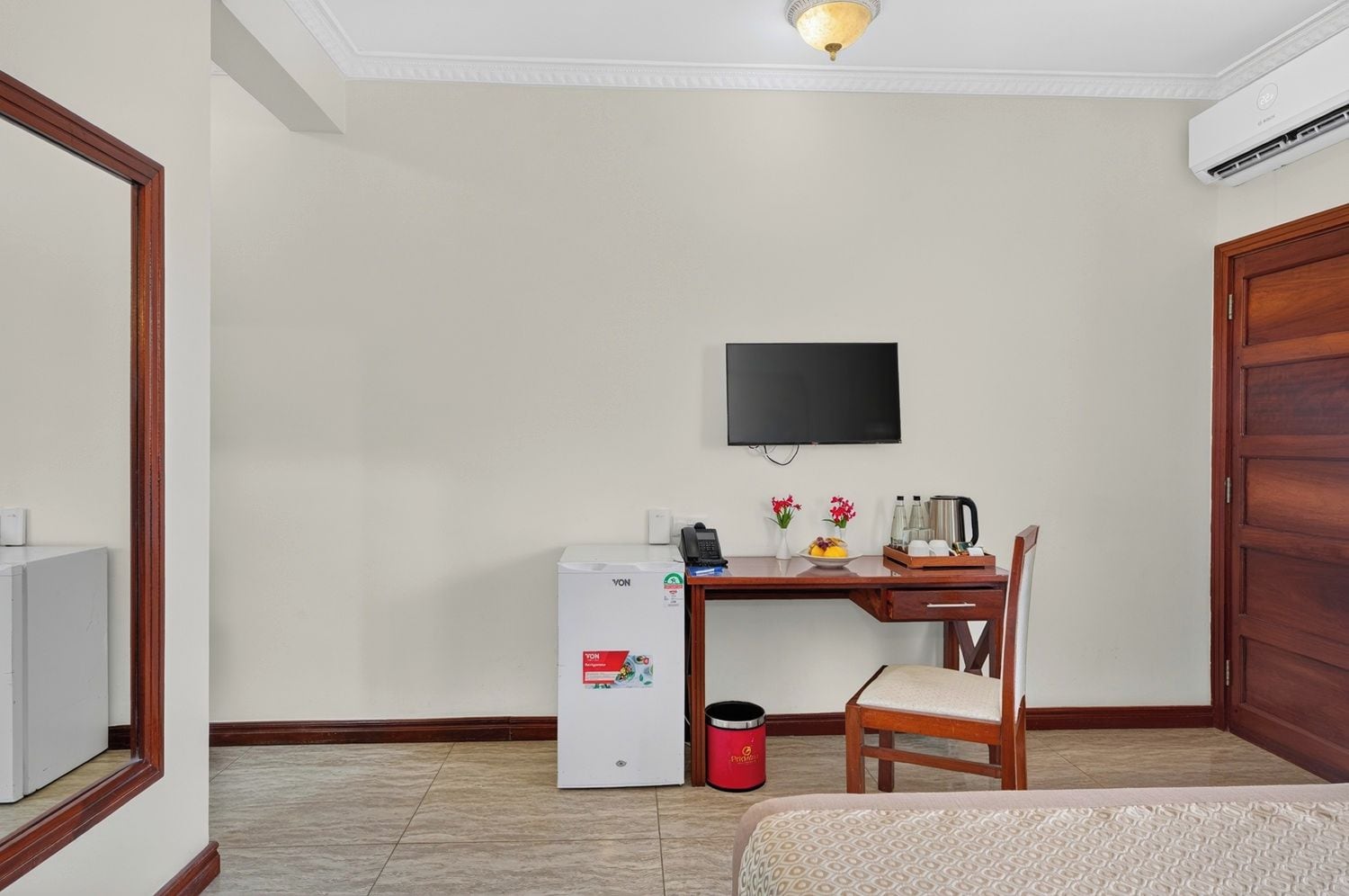 A hotel room with a bed, a desk with a chair, a television, a small refrigerator, and a trash bin.