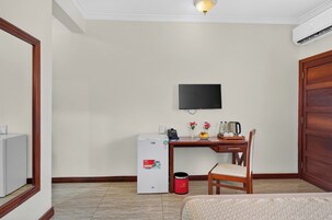 A hotel room with a bed, a desk with a chair, a television, a small refrigerator, and a trash bin.