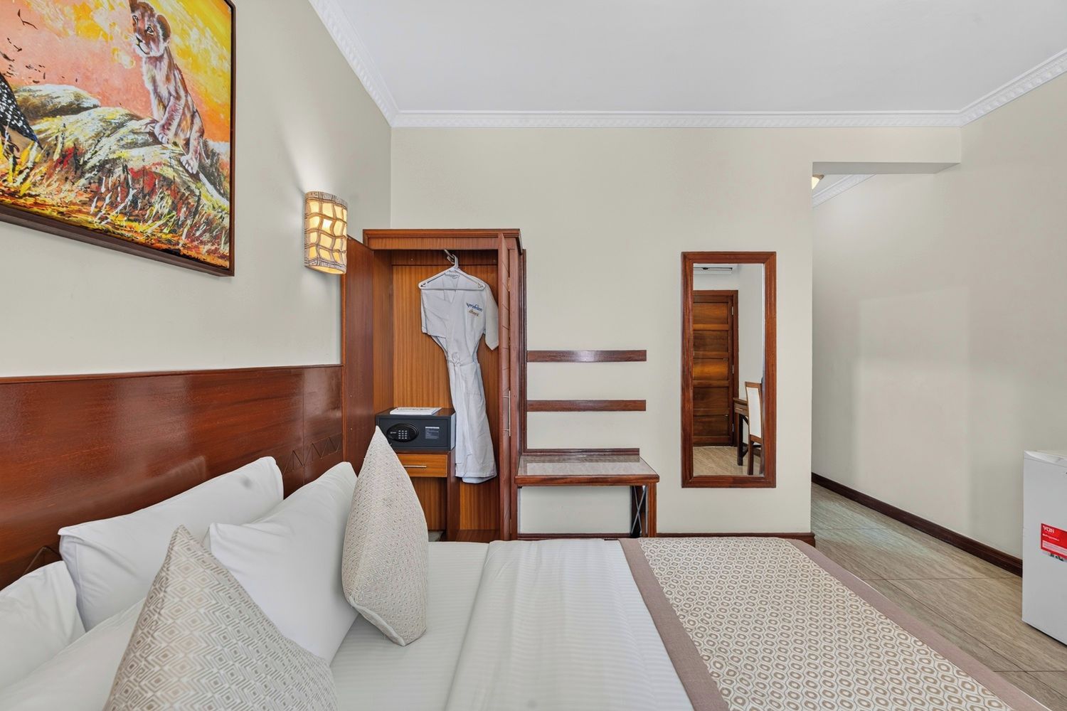 A hotel room with a bed, a bedside table, a robe hanging on a door, a wall-mounted lamp, and a painting.