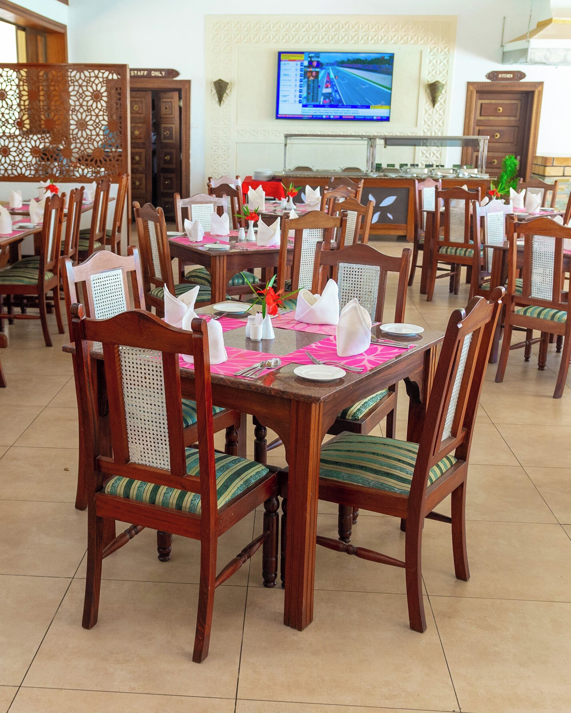 A dining area with wooden tables and chairs, a TV mounted on the wall, and a kitchen door in the background.