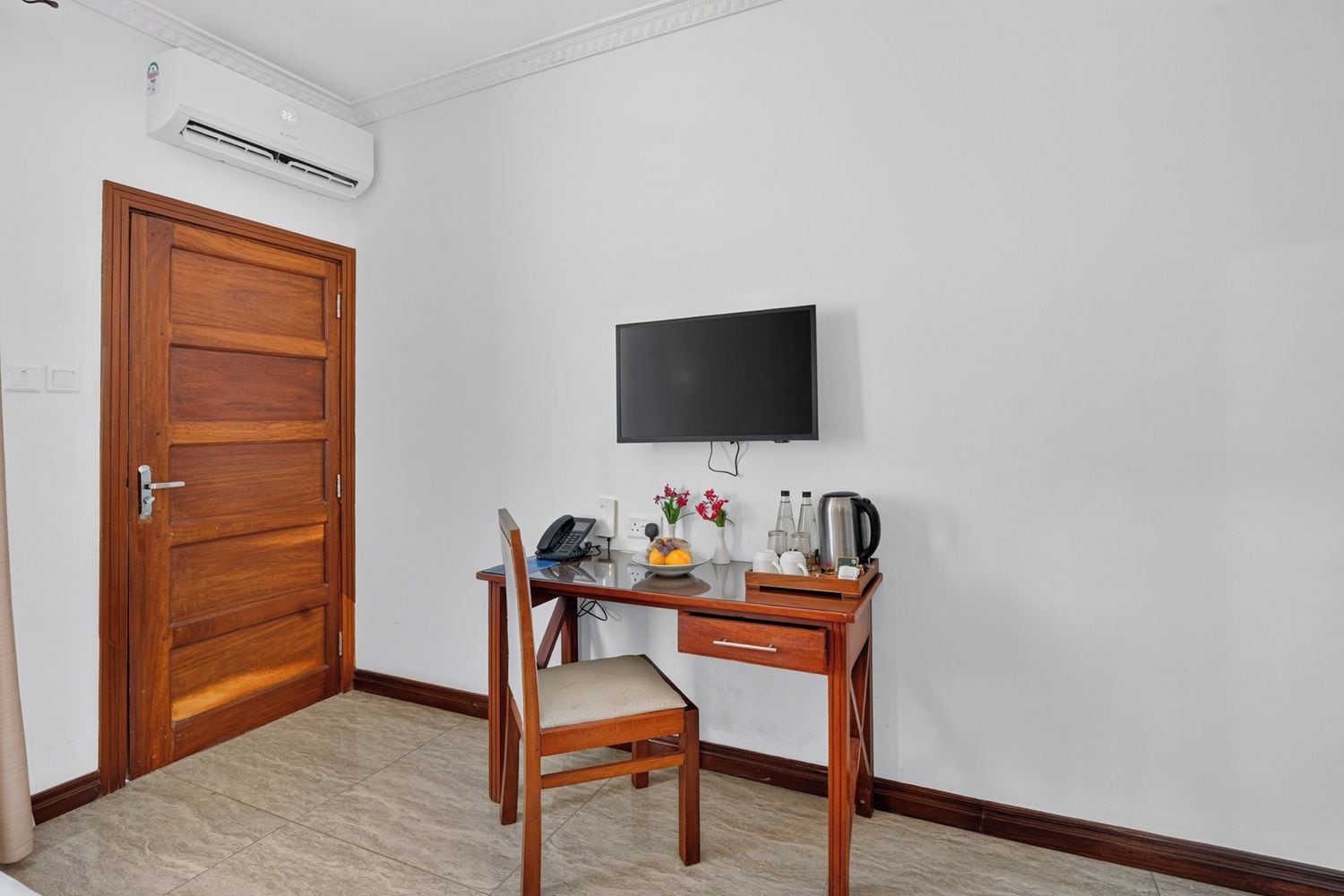 Une pièce avec une porte en bois, un bureau avec une chaise, un téléviseur à écran plat et une petite table sur laquelle se trouvent divers objets.