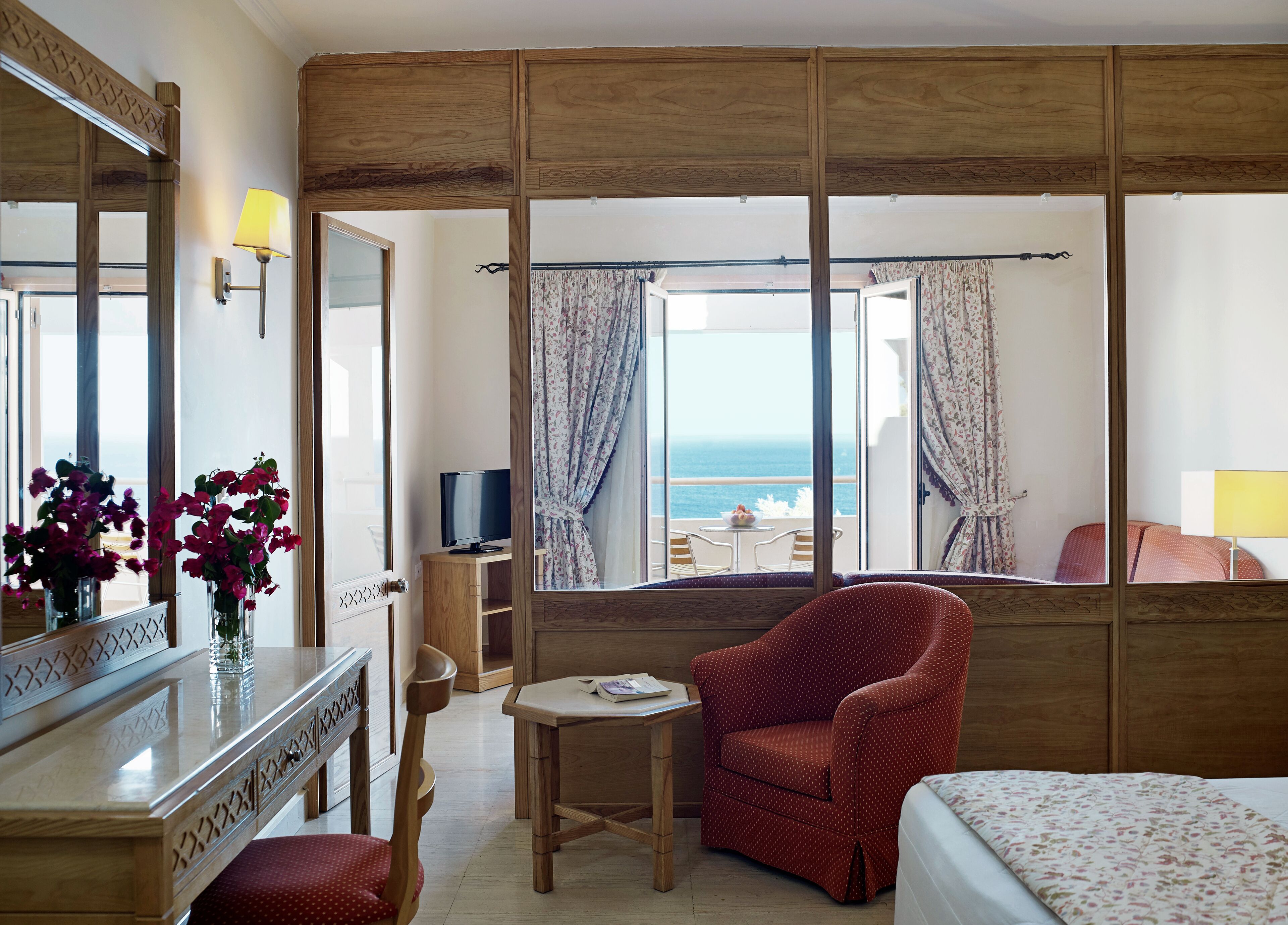 A hotel room with a wooden partition, a bed, a red polka dot chair, a small table with a book, a TV, and a view of the sea through the window.