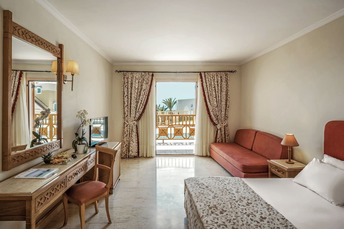 A hotel room with a bed, a desk with a chair, a dresser, a nightstand with a lamp, and a large mirror.