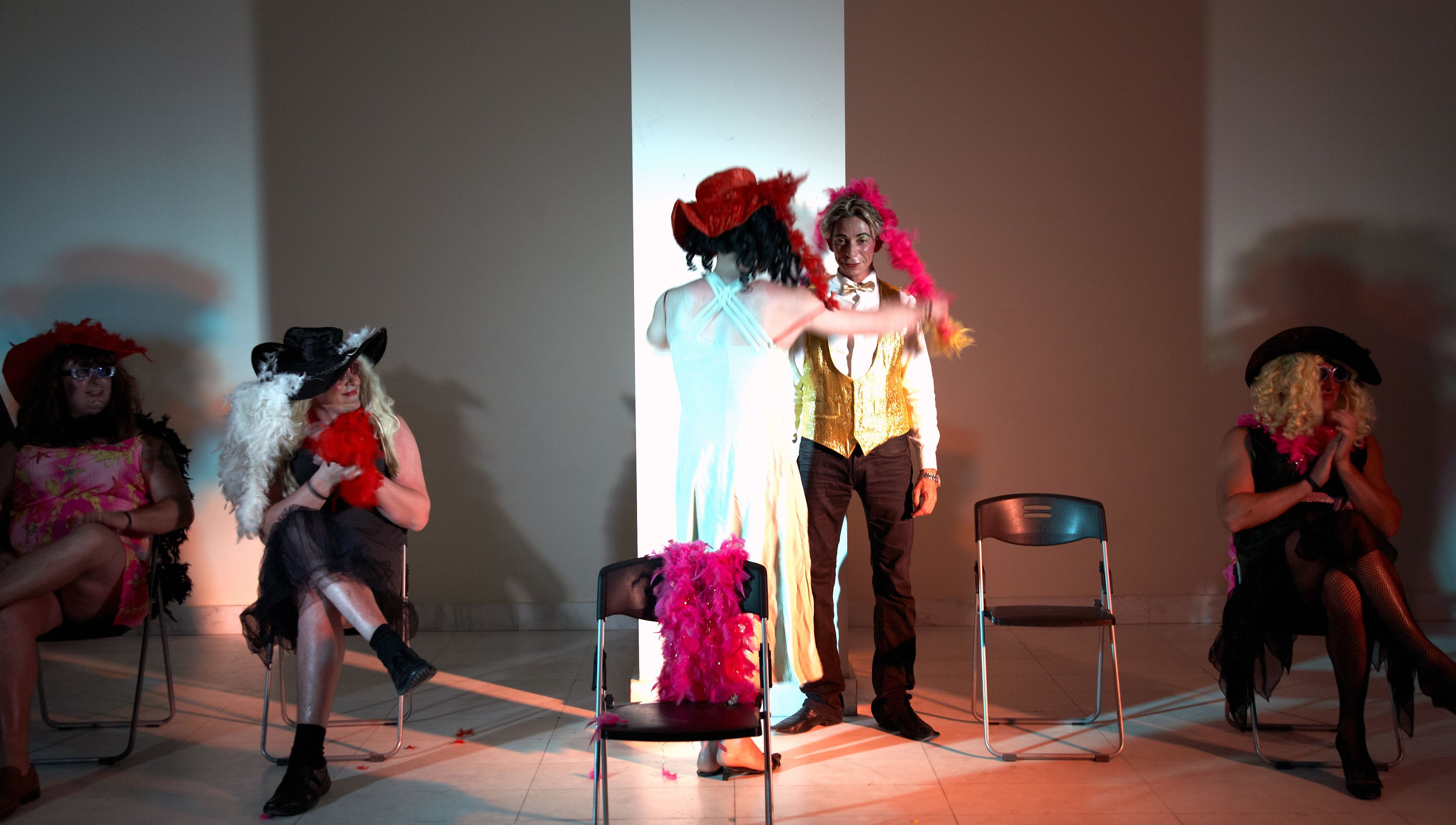A group of people in costumes seated on chairs in a room with a plain wall and a column.