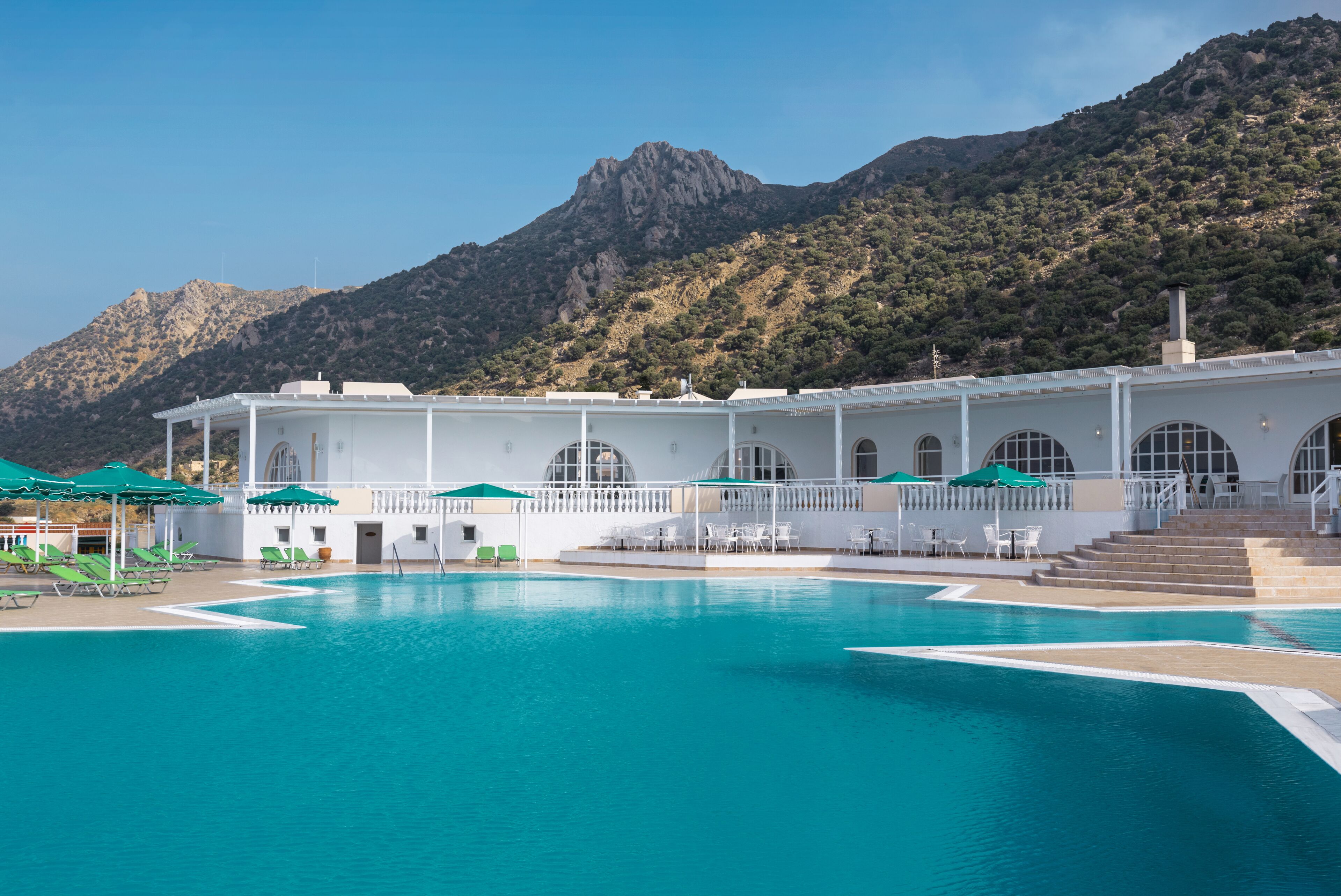 Ein Poolbereich mit Blick auf die Berge, ein weißes Gebäude mit Rundbogenfenstern und grünen Sonnenschirmen.