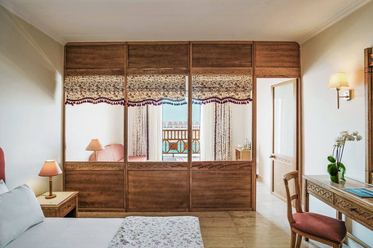 A bedroom with a wooden wardrobe, a bed with a floral bedspread, a nightstand with a lamp, a chair, and a desk with a vase of flowers.