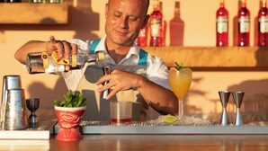 A bartender pouring a drink into a glass, with a bar counter and various bottles in the background. - Canvas by Mitsis Family Village (Kos)