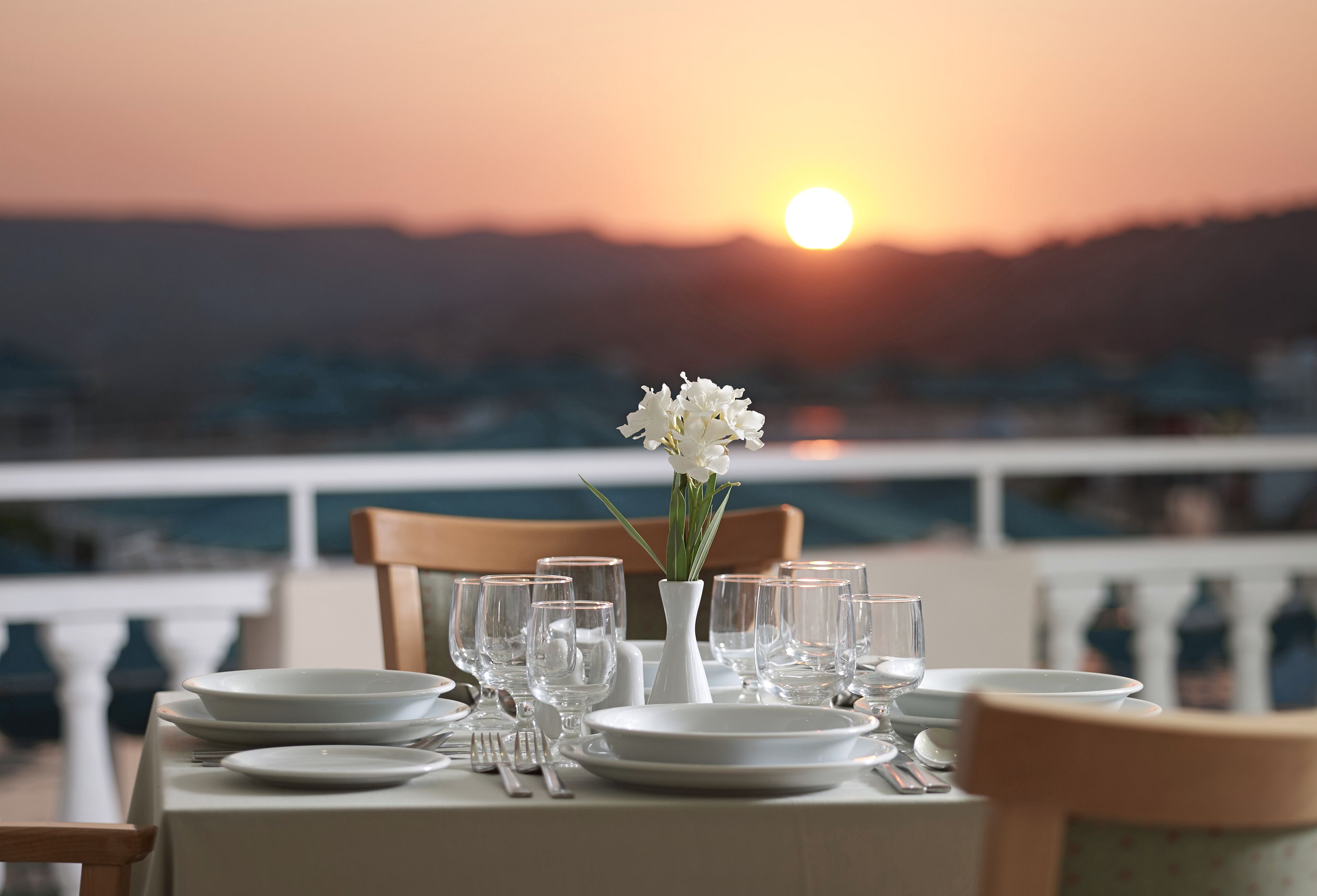 A table set for two with a vase of white flowers, glasses, and plates, overlooking a scenic sunset view.